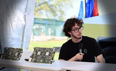 Koby Pierce on the panel at Chattanooga Charge, holding a microphone with Tesla HW3 and HW4 Full Self-Driving computers standing on the table beside him